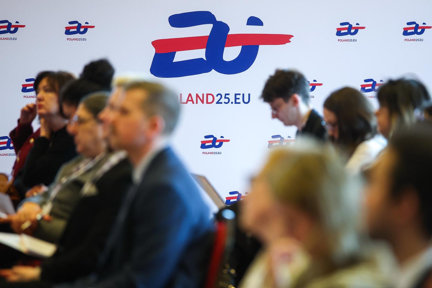 „Uczestnicy konferencji siedzący w sali, na tle baneru z logo ‘POLAND25.EU’ upamiętniającego 25-lecie członkostwa Polski w Unii Europejskiej.”