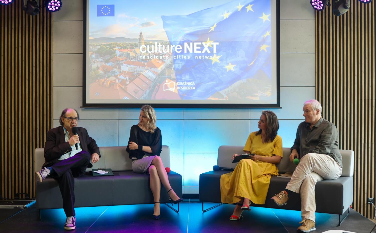 Na zdjęciu czterech ekspertów siedzi na scenie podczas panelu dyskusyjnego w nowoczesnej sali konferencyjnej. W tle znajduje się duży ekran wyświetlający grafikę z flagą Unii Europejskiej oraz napisem „culture NEXT – candidate cities network” i logiem Książnicy Beskidzkiej. Panelista po lewej stronie trzyma mikrofon i mówi, a pozostali uczestnicy słuchają go z zainteresowaniem. Oświetlenie sceniczne dodaje dynamiki wydarzeniu.