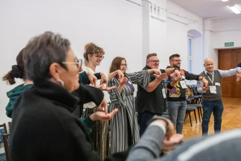 Na zdjęciu grupa osób uczestniczy w warsztatach z Humoru prowadzonych przez Czerwone Noski. Uczestnicy stoją w kręgu w jasnym pomieszczeniu, wykonując synchronizowane gesty dłońmi. Wszyscy wyglądają na zaangażowanych i uśmiechniętych, co tworzy atmosferę radości i współpracy.