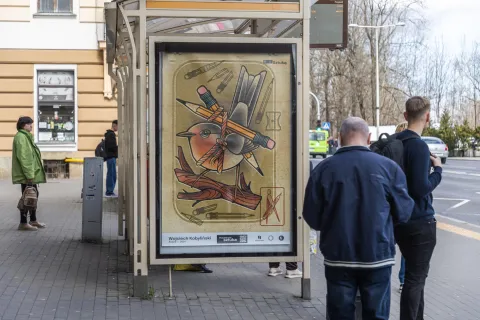 Ludzie czekają na przystanku autobusowym, obok którego znajduje się plakat z rysunkiem ptaka oplecionego ołówkami i kredkami. Ilustracja autorstwa Wojciecha Kobylińskiego jest częścią projektu „Przystanek Sztuka”.