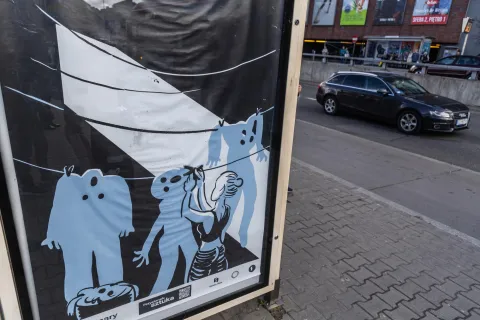 Plakat artystyczny na przystanku autobusowym przedstawia stylizowaną postać kobiety wieszającej na sznurku niebieskie ubrania przypominające ludzkie sylwetki. Scena oświetlona jest snopem światła, co nadaje kompozycji dramatyczny, symboliczny charakter. Obok plakatów przejeżdżają samochody, a w tle widać galerię handlową.