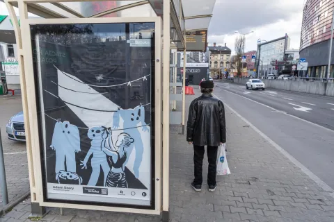 Mężczyzna w czarnej kurtce i czapce stoi na przystanku autobusowym, obok plakatu z projektu „Przystanek Sztuka”. Ilustracja autorstwa Adama Szarego zatytułowana „Suszarnia trupów” przedstawia postać wieszającą na sznurku puste, niebieskie sylwetki przypominające ludzkie ciała. W tle ruchliwa ulica z samochodami i zabudową miejską.