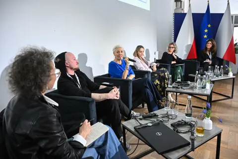 Na zdjęciu widzimy panel dyskusyjny z udziałem sześciu osób siedzących w półokręgu na scenie. Jedna z uczestniczek trzyma mikrofon i mówi, pozostali uważnie słuchają. Przed nimi znajdują się stoliki z butelkami wody, filiżankami, laptopami oraz dekoracyjnymi kwiatami. W tle stoją flagi Polski i Unii Europejskiej. Panuje swobodna, ale profesjonalna atmosfera, typowa dla konferencji lub debat eksperckich.