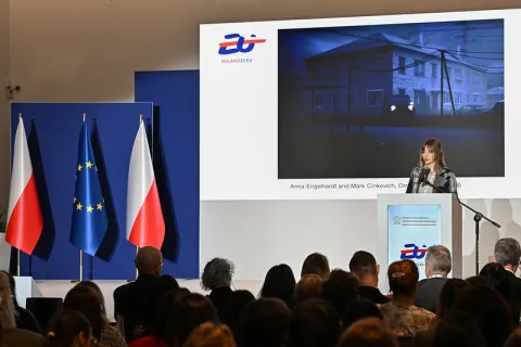 Na zdjęciu widzimy kobietę przemawiającą z mównicy podczas oficjalnego wydarzenia. Za nią znajduje się duży ekran prezentujący grafikę oraz logotyp „POLAND25.EU”. Na grafice widnieje podpis: „Anna Engelhardt and Mark Cinkevich, Onset (2021)”. W tle stoją cztery flagi – dwie polskie i dwie Unii Europejskiej. Na sali obecna jest publiczność, uważnie słuchająca wystąpienia. Mównica opatrzona jest logiem Ministerstwa Kultury i Dziedzictwa Narodowego oraz znakiem graficznym POLAND25.EU.