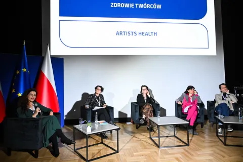 Na zdjęciu widzimy pięcioosobowy panel dyskusyjny zorganizowany w przestrzeni konferencyjnej. Uczestnicy siedzą na fotelach ustawionych w półokręgu, a za nimi wyświetlony jest tytuł dyskusji w dwóch językach: „ZDROWIE TWÓRCÓW / ARTISTS HEALTH”. Po lewej stronie sceny znajdują się flagi Unii Europejskiej i Polski. Przed panelistami ustawione są stoliki z wodą i drobnymi dekoracjami. Jedna z osób mówi do mikrofonu, pozostali przysłuchują się uważnie. Panuje profesjonalna atmosfera spotkania poświęconego kondy
