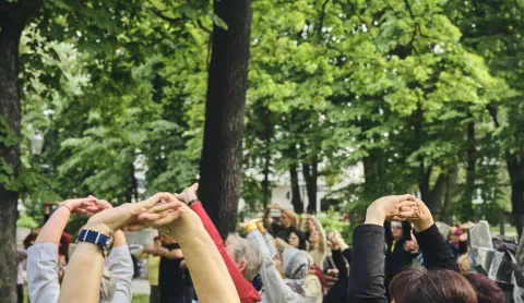Grupa ludzi ćwiczących na świeżym powietrzu w parku, unoszących ręce do góry w geście rozciągania. W tle zielone drzewa tworzą przyjemną, naturalną atmosferę.