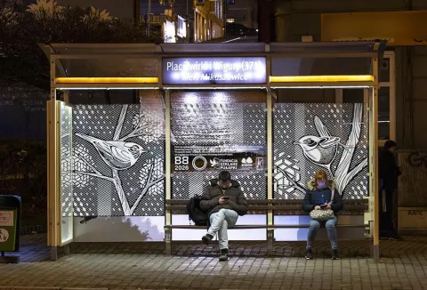 Wieczorna scena na przystanku autobusowym "Plac Żwirki i Wigury" w Bielsku-Białej. Na szklanych panelach wiaty widnieją czarno-białe grafiki ptaków na gałęziach, będące częścią projektu artystycznego. Dwoje pasażerów siedzi na ławce, skupionych na telefonach. Światła miasta i podświetlony szyld przystanku nadają atmosferę nocnej przestrzeni miejskiej.