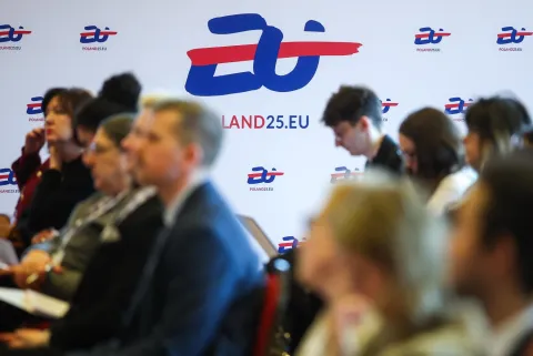 „Uczestnicy konferencji siedzący w sali, na tle baneru z logo ‘POLAND25.EU’ upamiętniającego 25-lecie członkostwa Polski w Unii Europejskiej.”