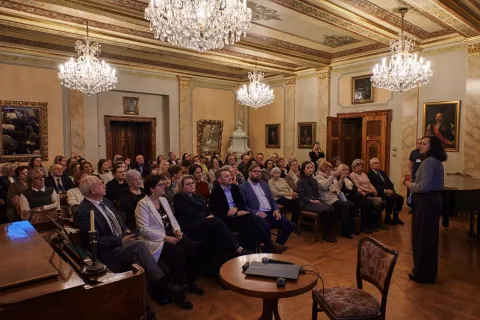 W eleganckiej, zabytkowej sali pałacowej odbywa się wykład o muzeoterapii. Prelegentka stoi przy stole z laptopem i mikrofonem, zwracając się do licznej publiczności, która uważnie słucha, robi notatki i fotografuje. Kryształowe żyrandole, historyczne portrety i bogate zdobienia tworzą atmosferę intelektualnej wymiany myśli w otoczeniu dziedzictwa kulturowego.